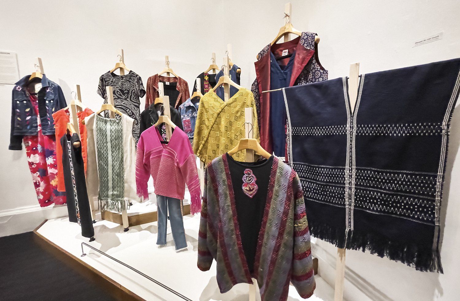 Clothing display with colorful patterned garments and shawls hanging on a white wall in a gallery