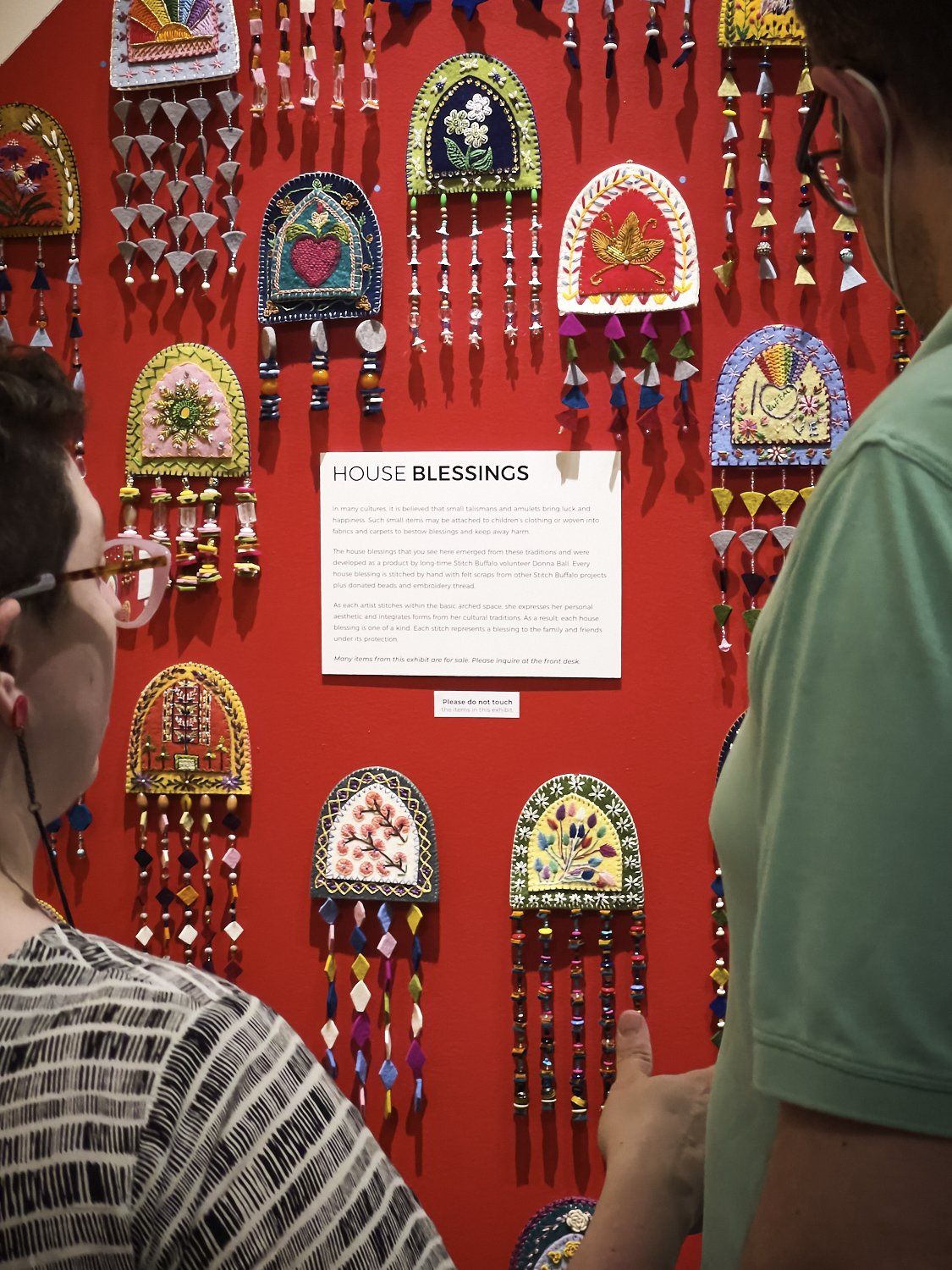 Red wall display of colorful hanging ornaments with a central informational plaque; two visitors at the edges