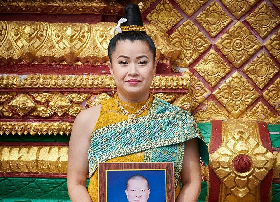 Woman in a gold and teal traditional dress holding a framed portrait, standing before a gilded patterned backdrop