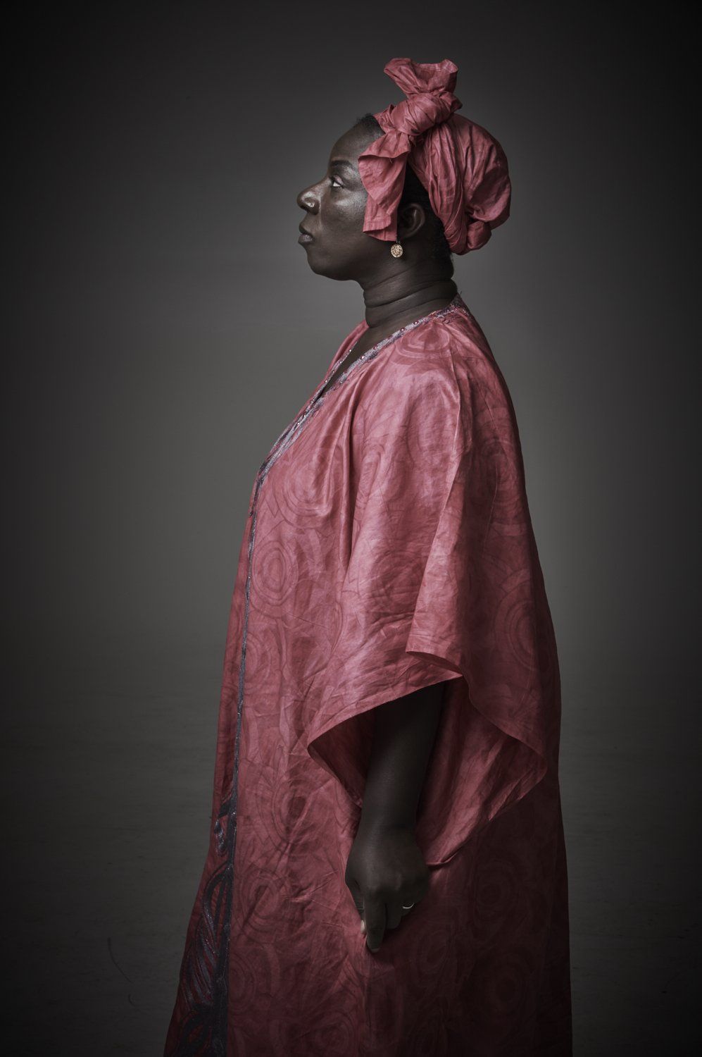 Side profile of a person in a pink textured garment and headwrap against a dark studio backdrop