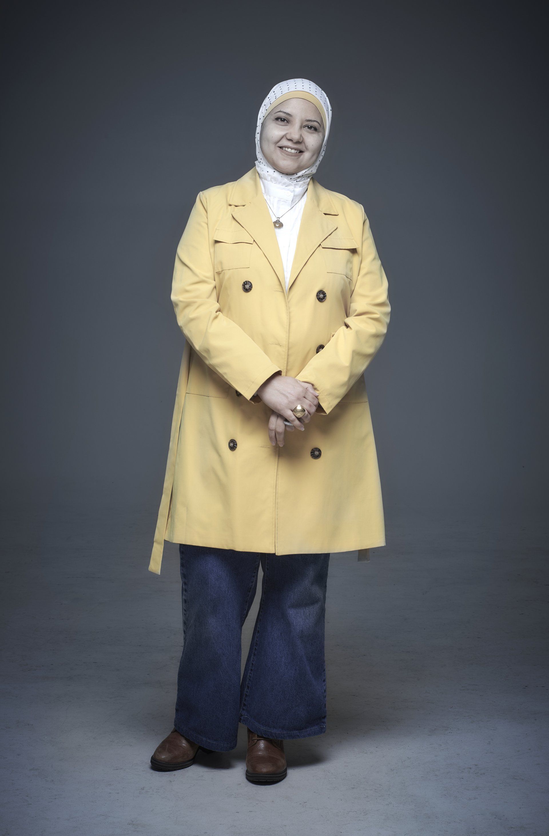 Person standing against a gray backdrop, wearing a yellow coat, white headscarf, and jeans.