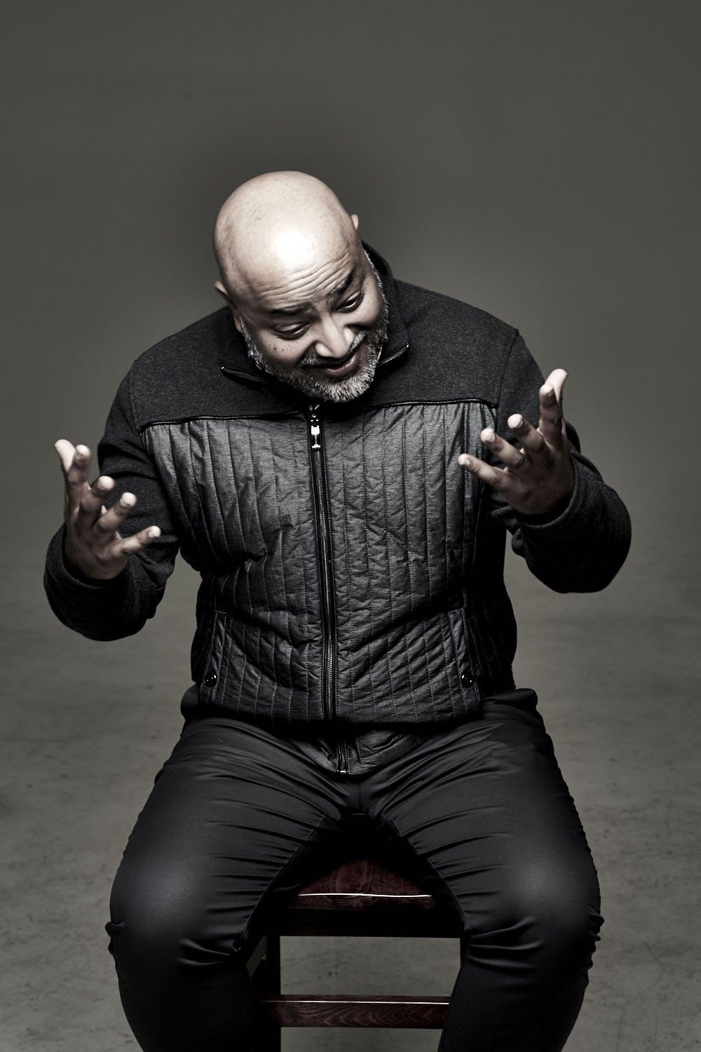 Man in dark clothing seated against a gray backdrop, gesturing with both hands and looking downward.