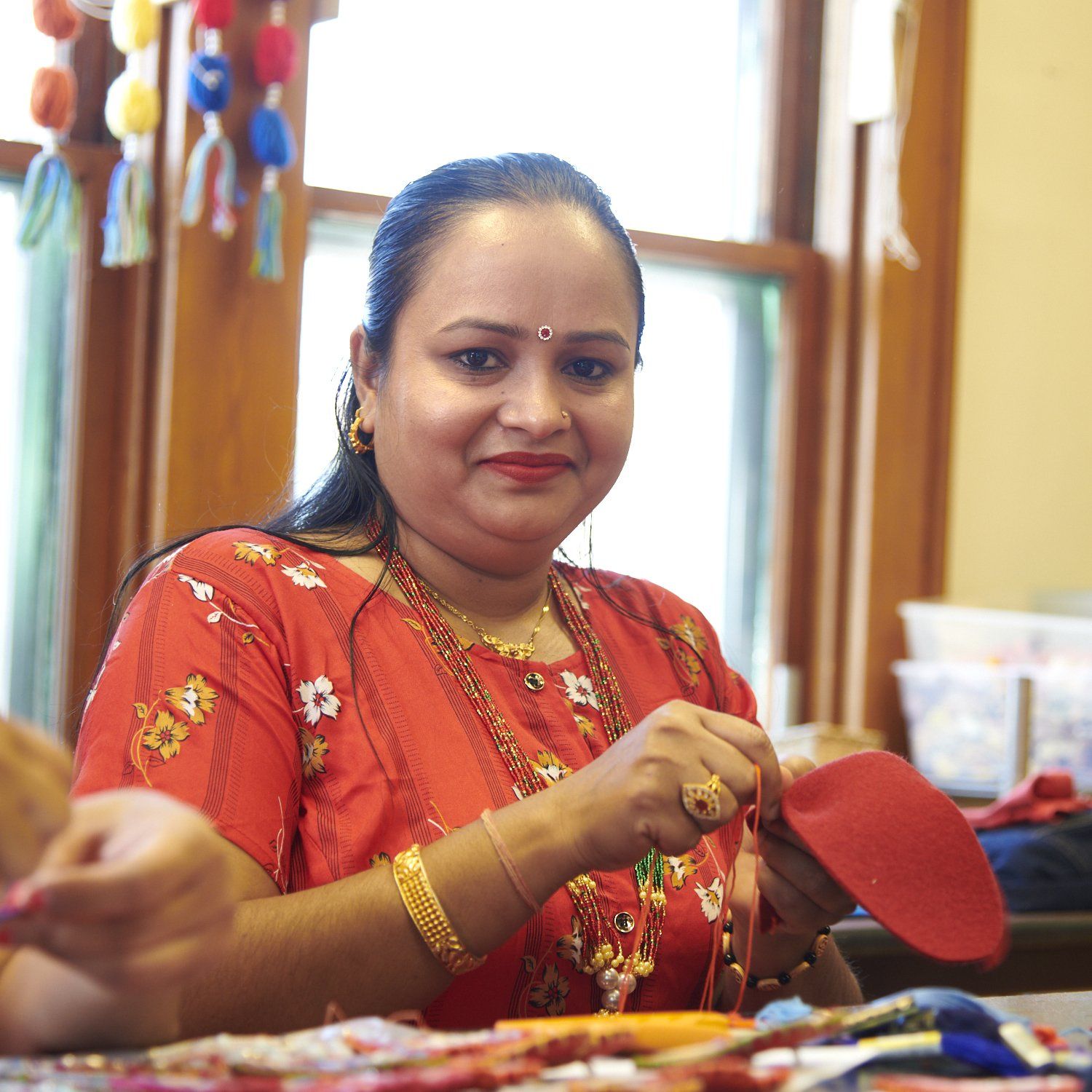 Woman in an orange blouse and gold jewelry smiling while stitching fabric at a sewing table