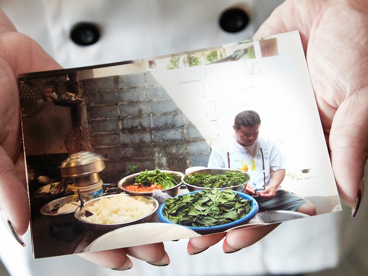 Person holding a small photo of a meal with greens, rice, and a seated cook at a table