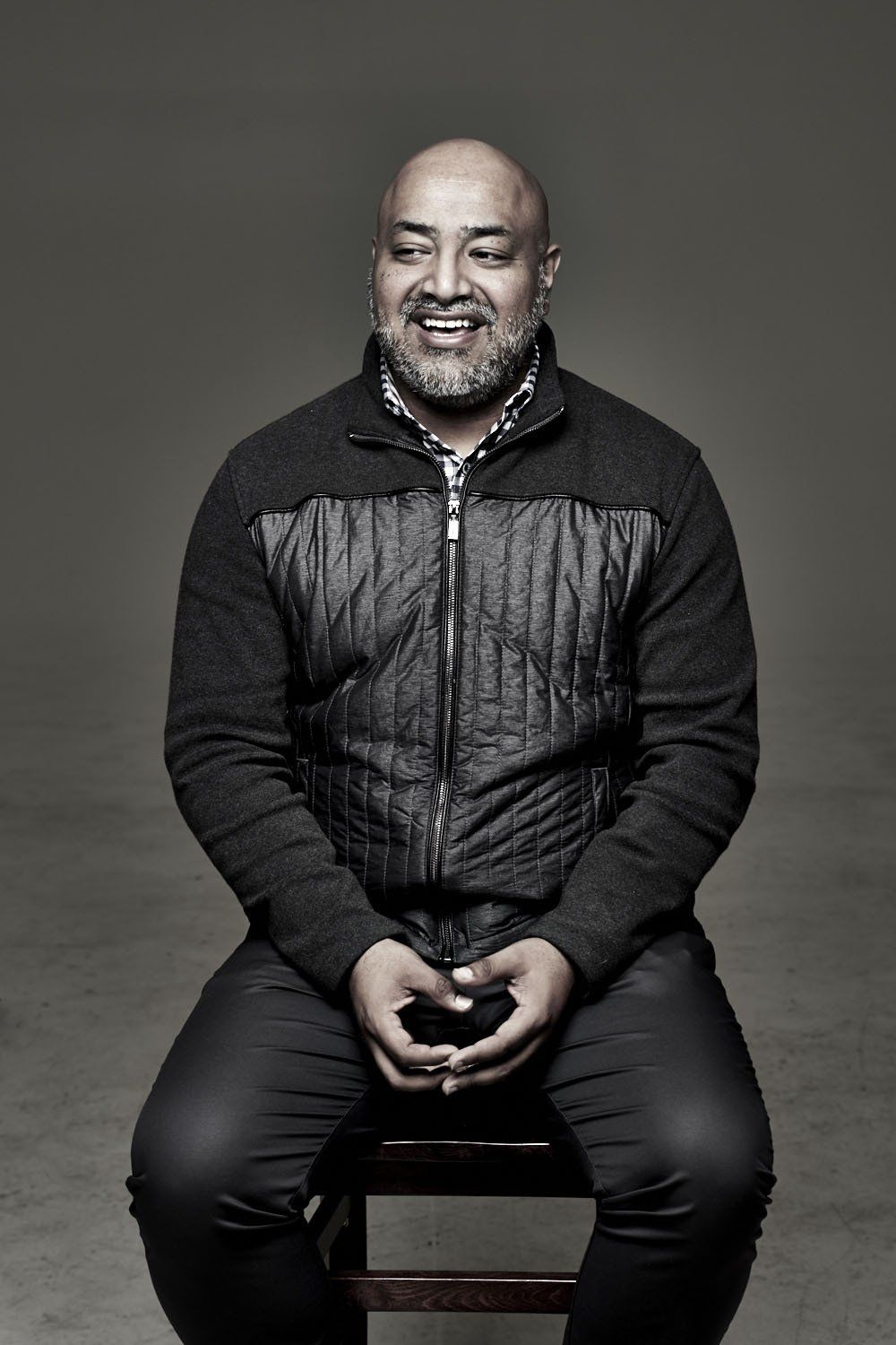 Smiling man in a dark jacket sits on a stool against a gray studio backdrop.