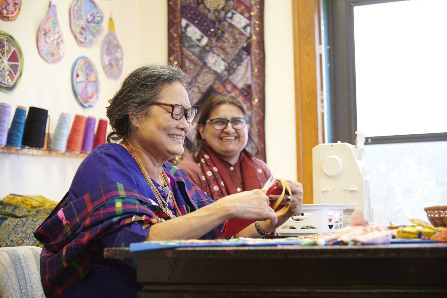 Two people smiling at a craft table, holding sewing materials in a bright workshop