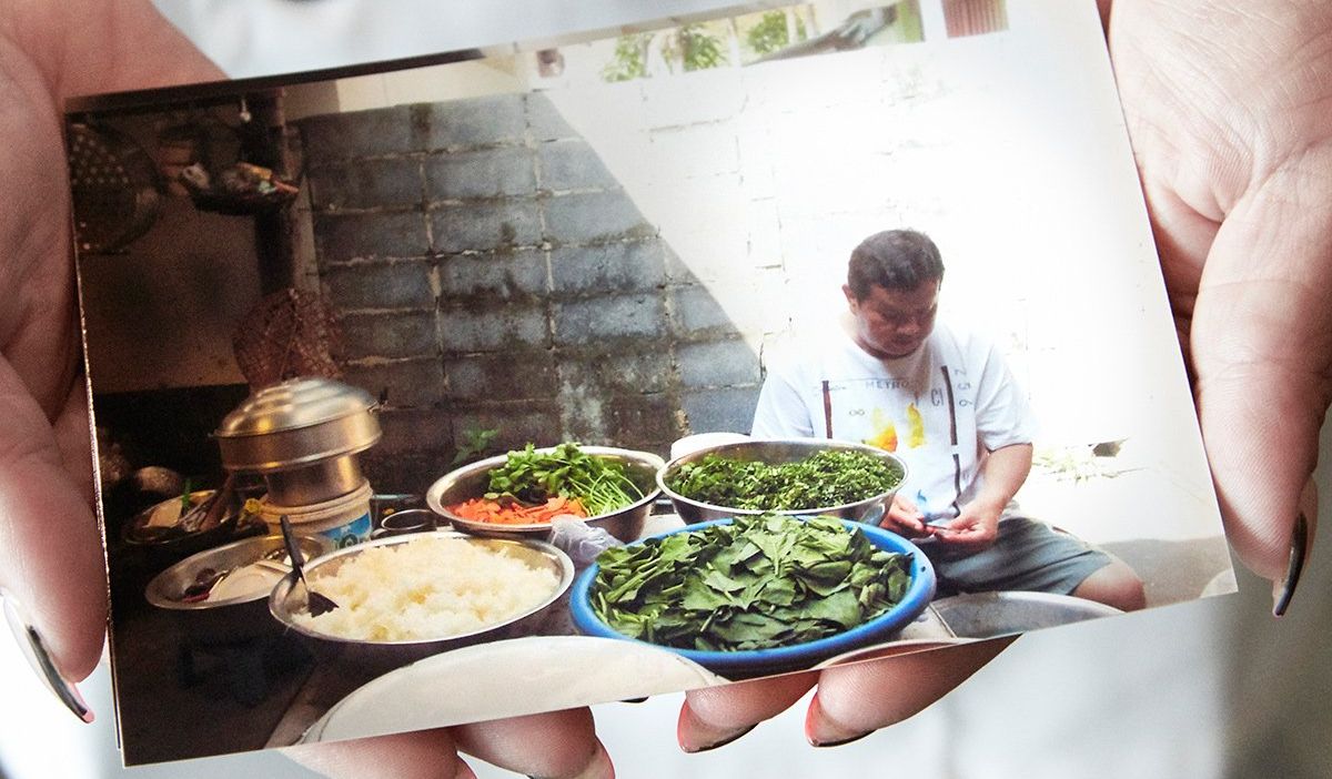 A hand holds a photo of a man seated beside plates of food at an outdoor table.