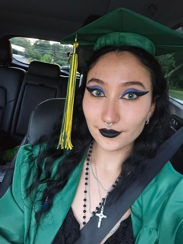 Person in green graduation cap and gown with dark makeup, seated in a car and wearing a seatbelt.