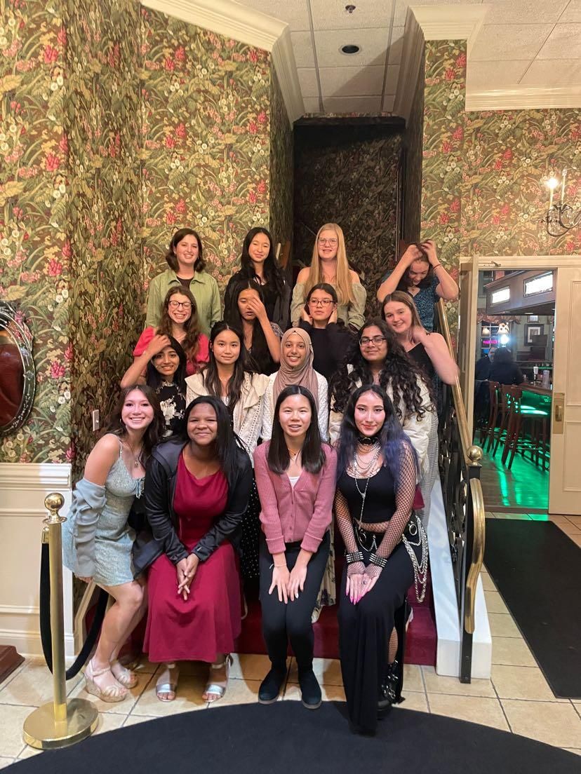 Group posing on a staircase indoors, smiling and making heart gestures, with patterned wallpaper and warm lighting.