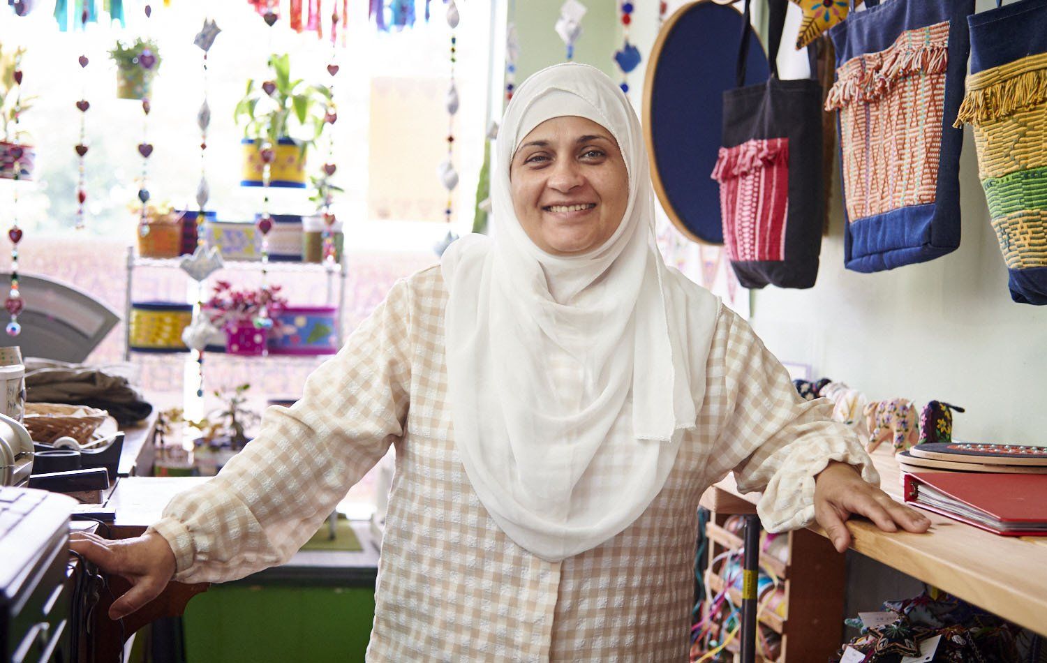 Shopkeeper smiling behind a counter in a colorful market stall with hanging baskets and crafts