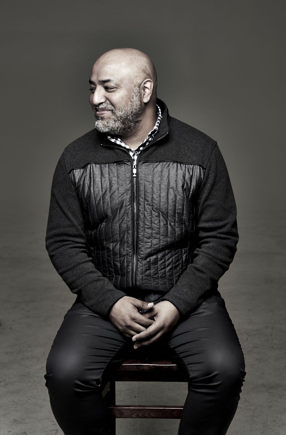 Man seated on a stool against a gray backdrop, wearing a black jacket and looking to the side.