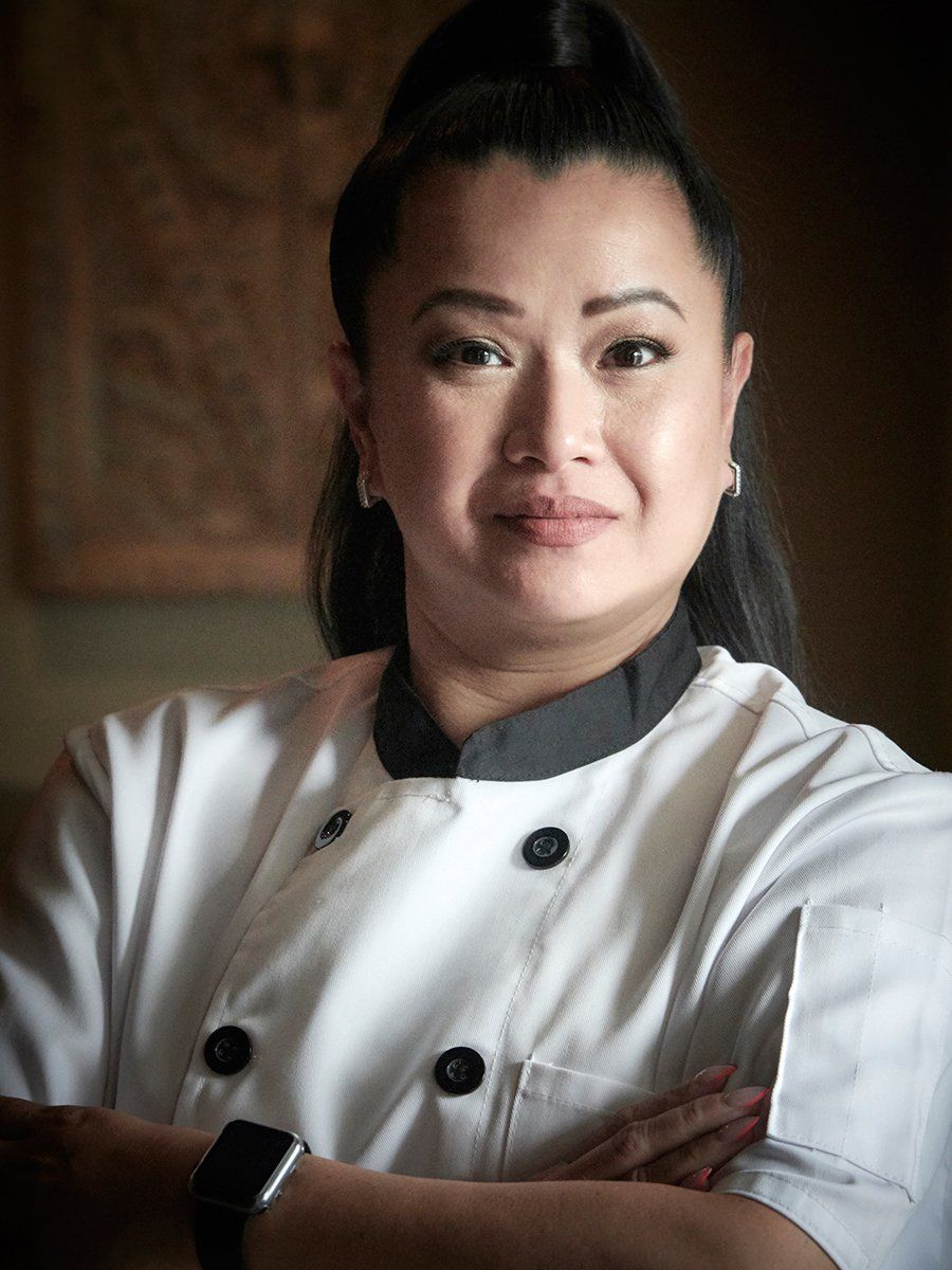 Chef in a white jacket with arms crossed, standing indoors with a calm expression.