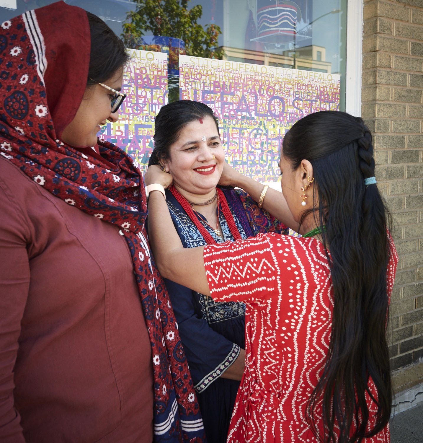 Three women smiling and talking outdoors near a brick wall and colorful signboard.
