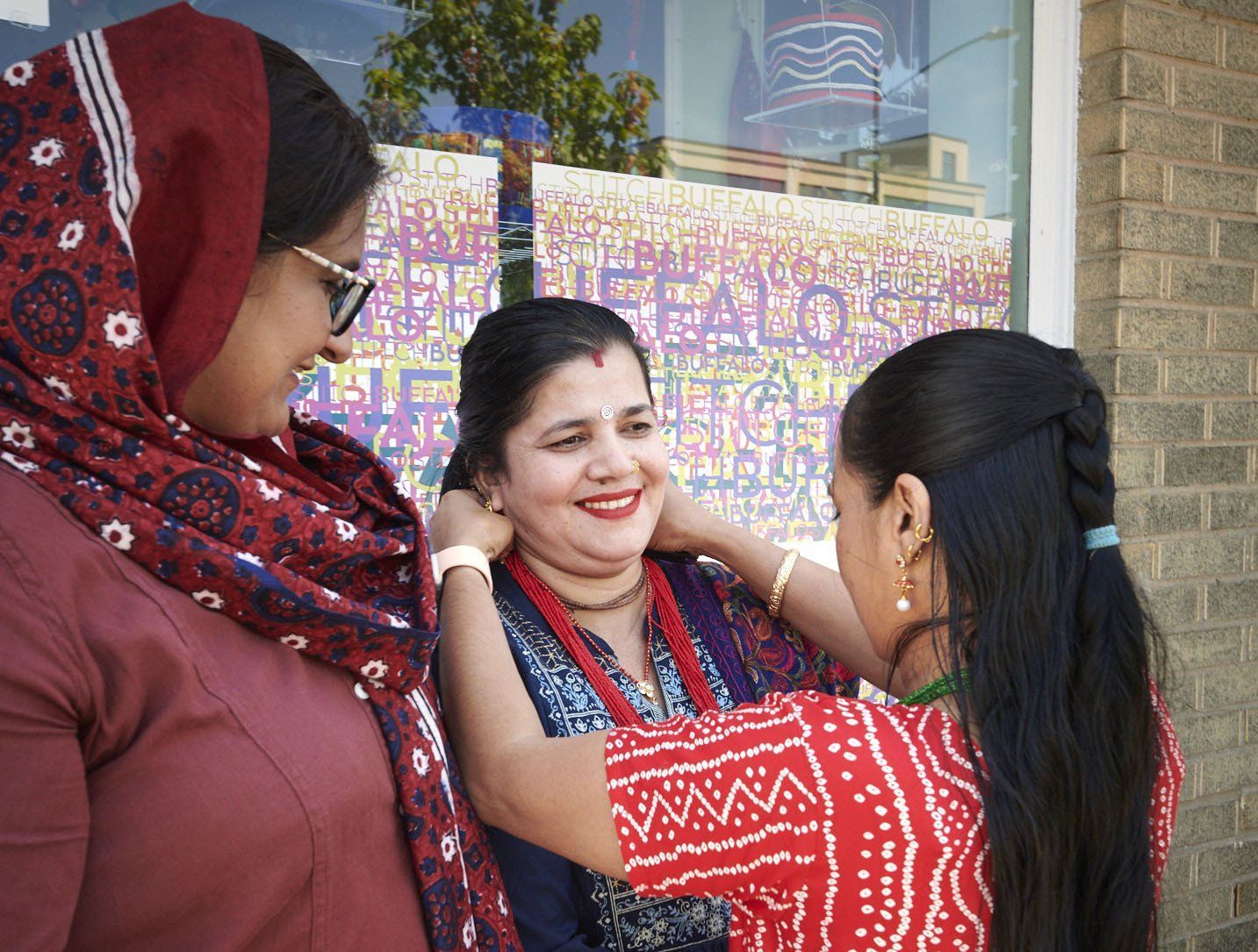 Three people chatting outdoors, with one smiling in a red patterned outfit being adjusted by another.