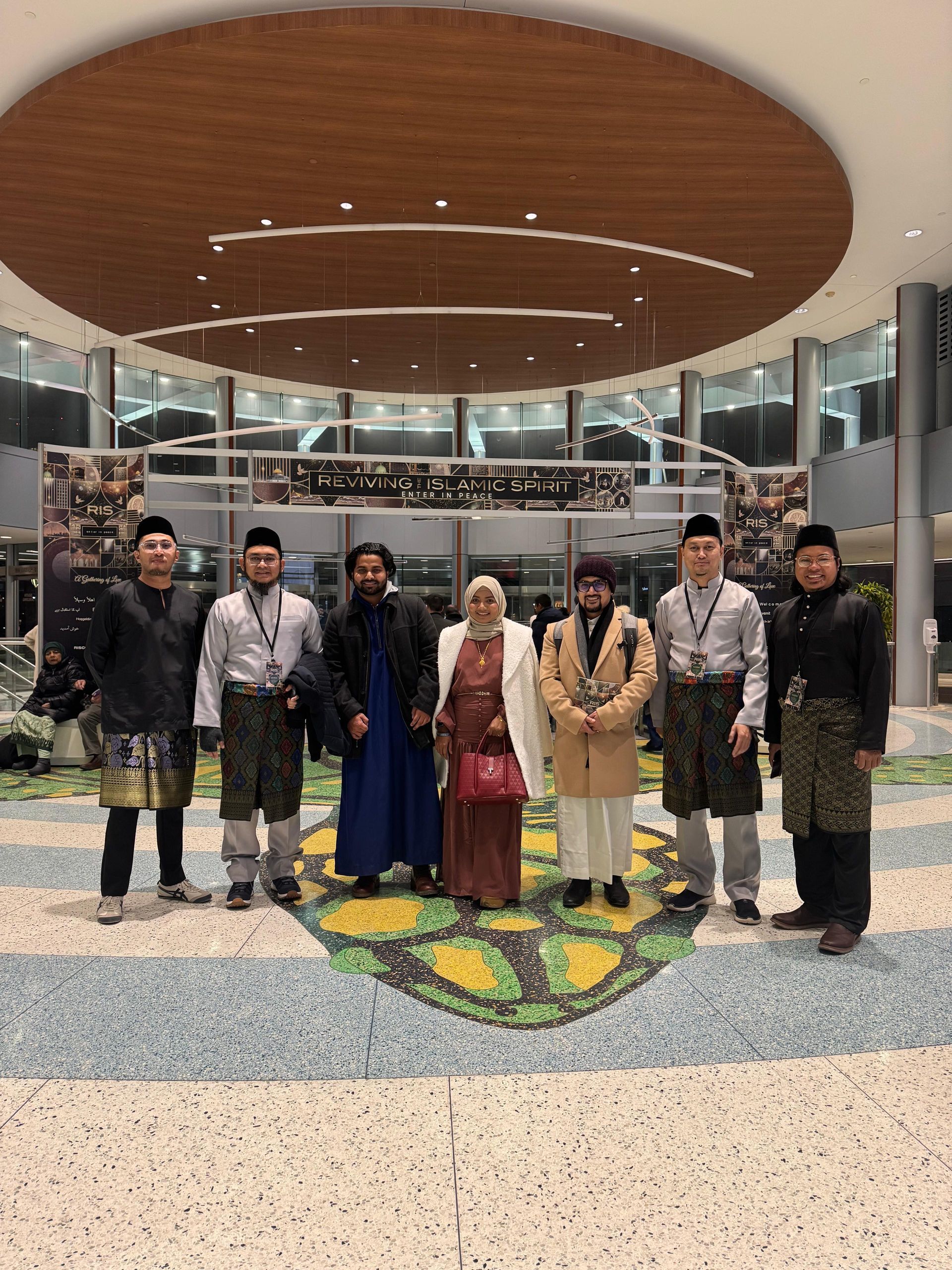 Seven people posing in a modern lobby beneath a large oval ceiling feature.