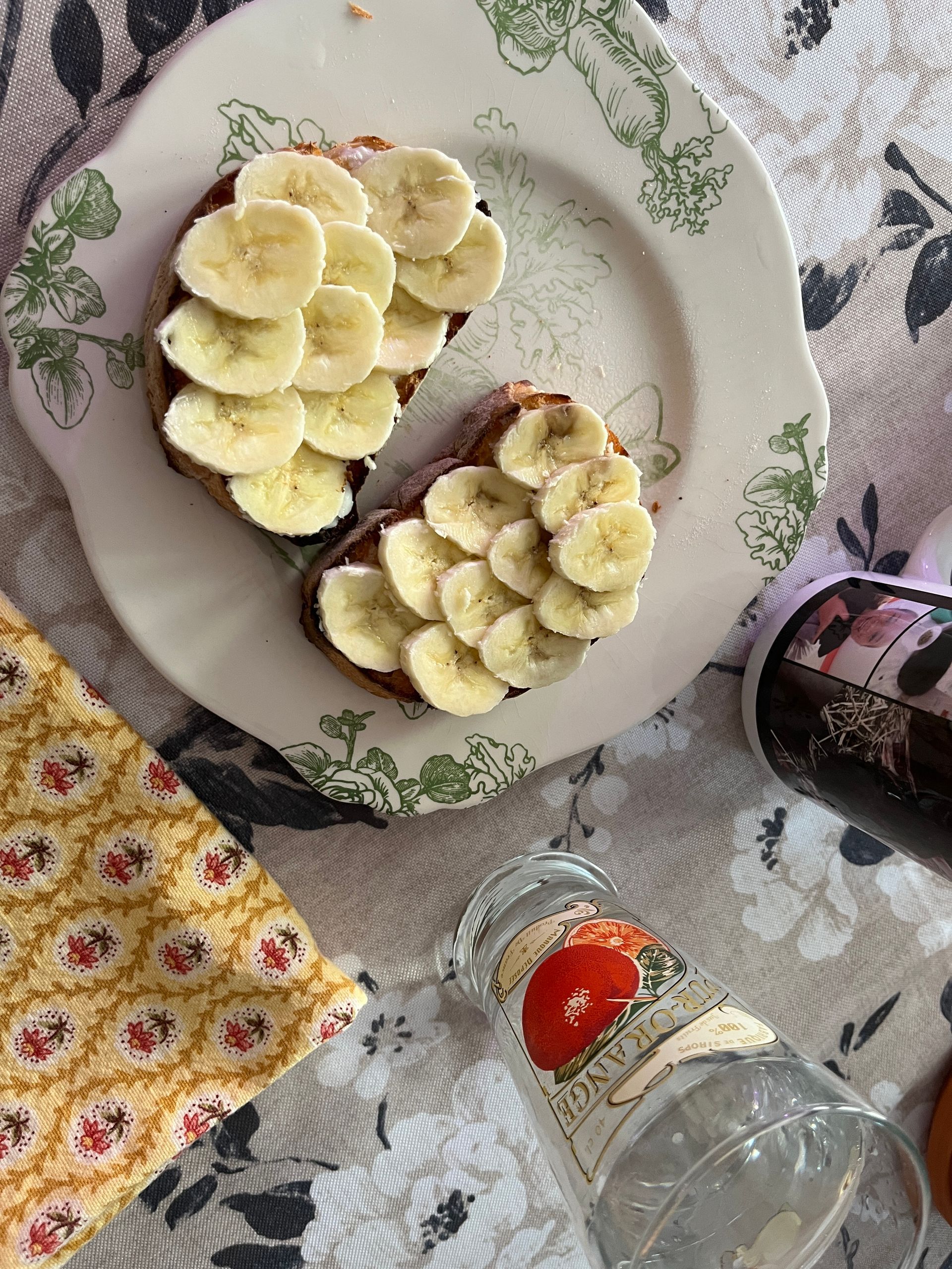Banana slices on toast on a patterned plate beside a tipped glass bottle and cloth napkin