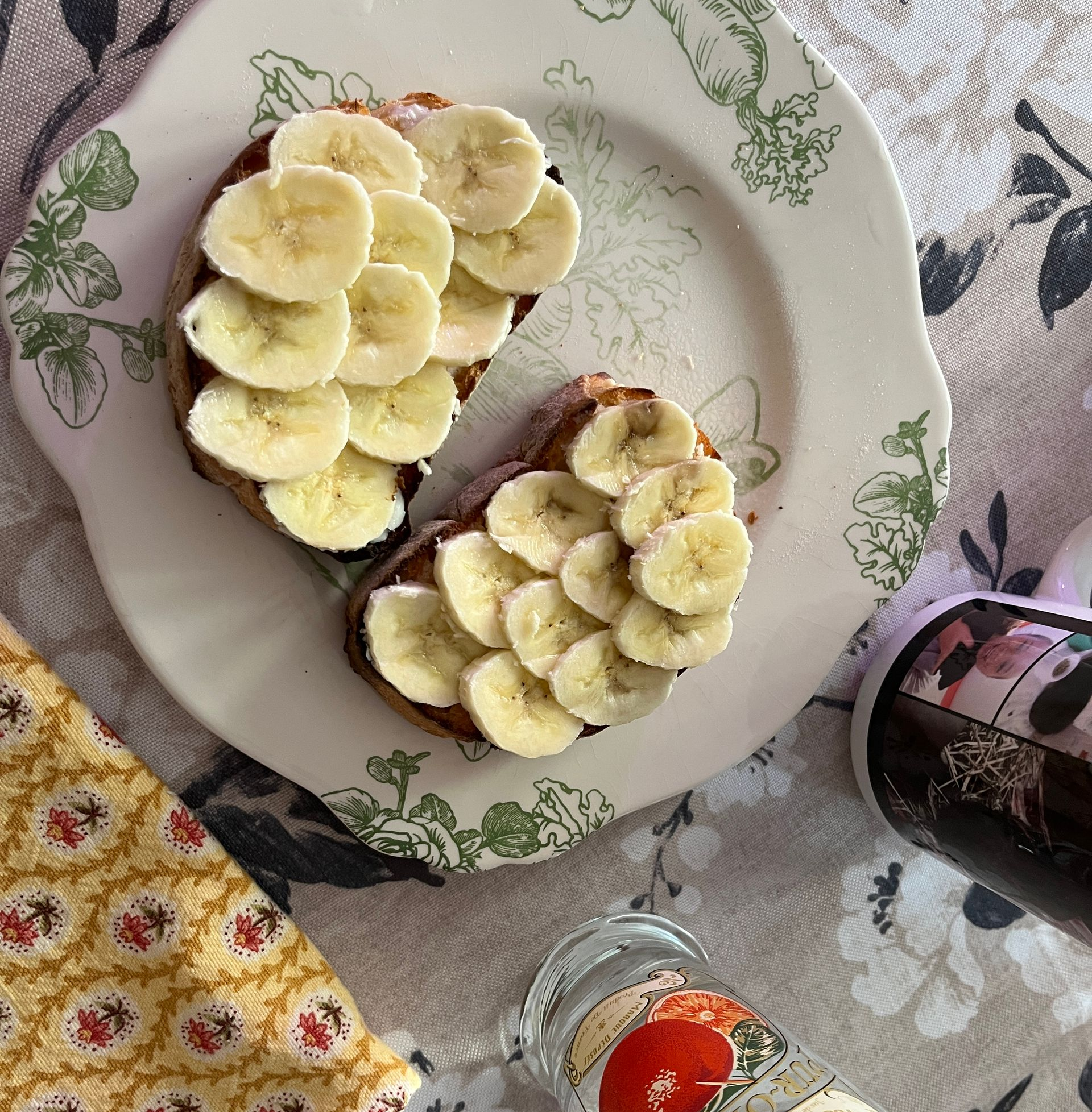 Plate with banana-topped toast on a floral tablecloth, with a small jar and fabric napkin nearby