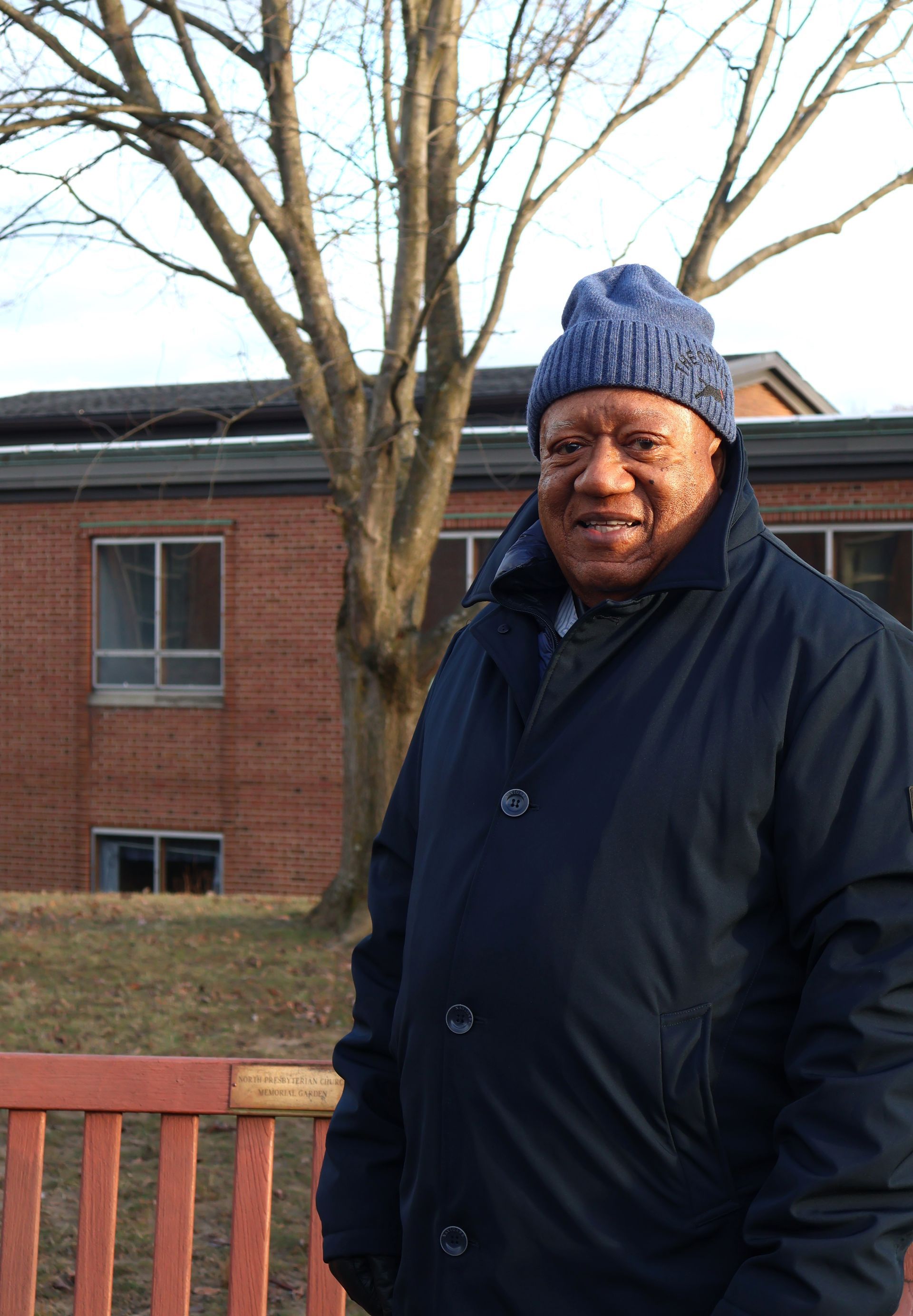 Man standing outdoors by a red railing, wearing a dark coat and blue knit hat, with leafless trees and a brick building behind him.