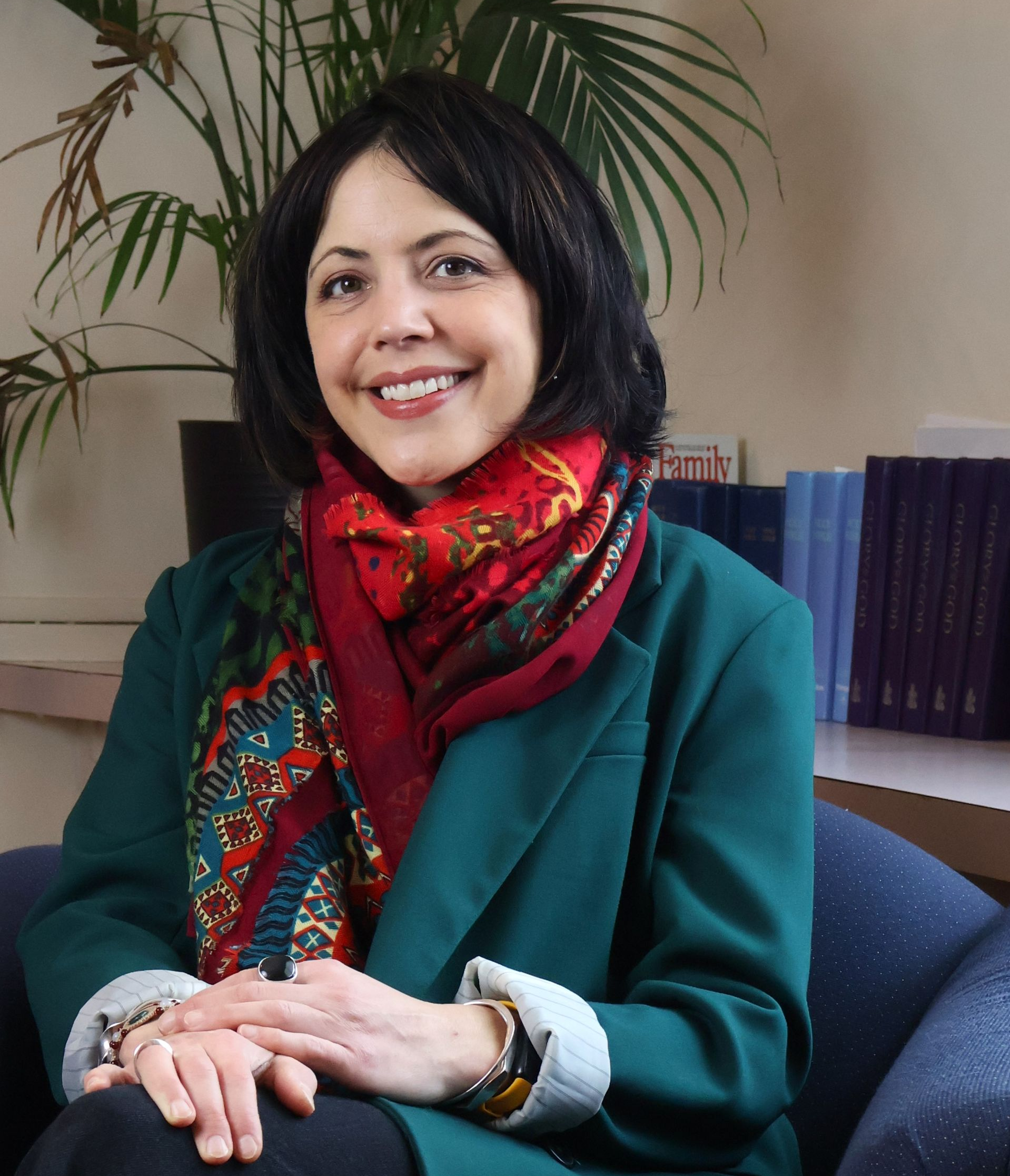 Woman seated indoors, smiling, wearing a green coat and red scarf, with bookshelves and a plant behind her.
