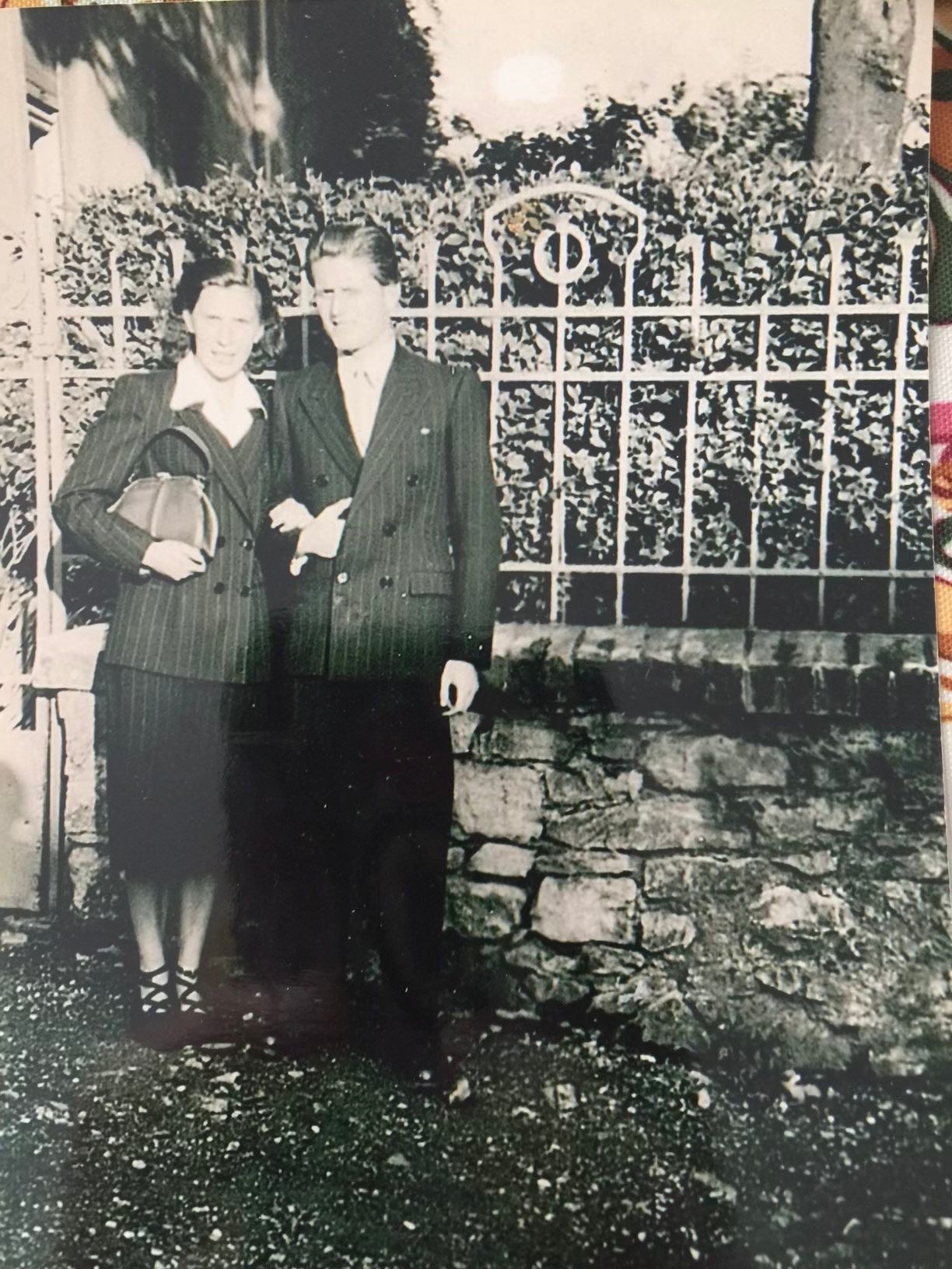 Two people standing together by a fence in front of a stone wall, in a vintage black-and-white photo.
