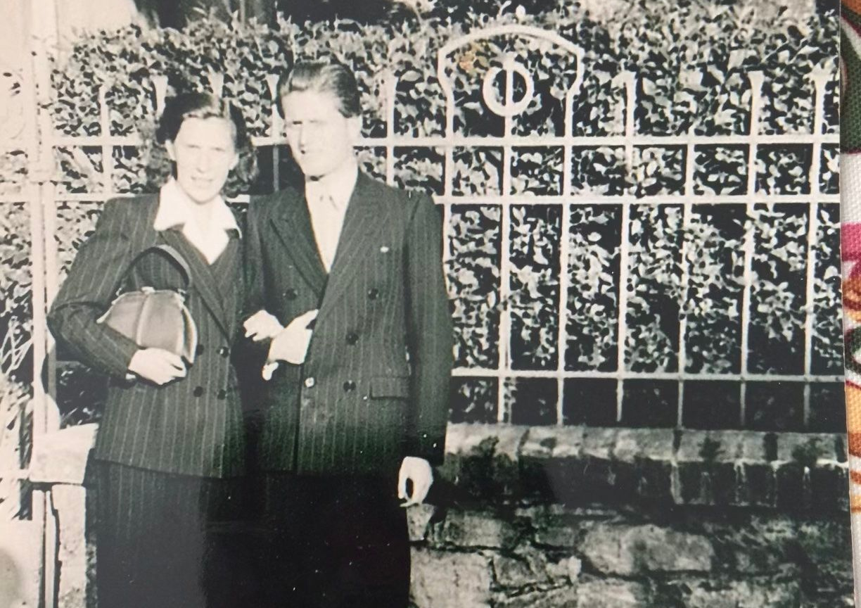 Two people standing together in front of a decorative iron fence, one holding a handbag