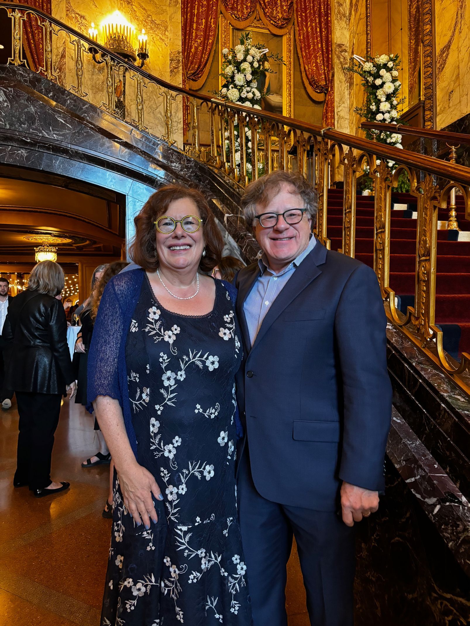 Two people smiling in formal attire inside an ornate, gold-lit hall.