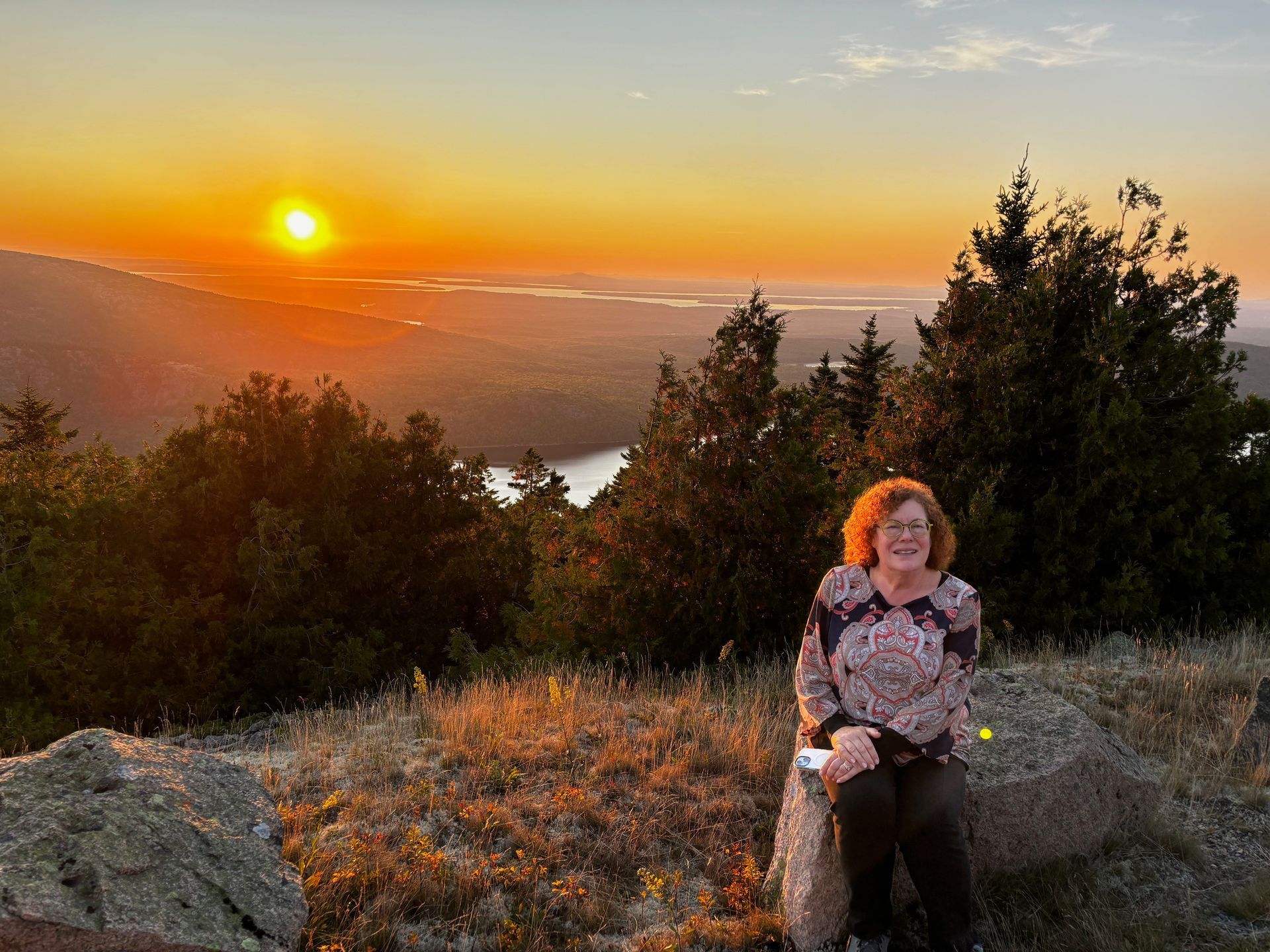 Woman sitting on a rock at sunset in a mountain overlook with trees and orange sky