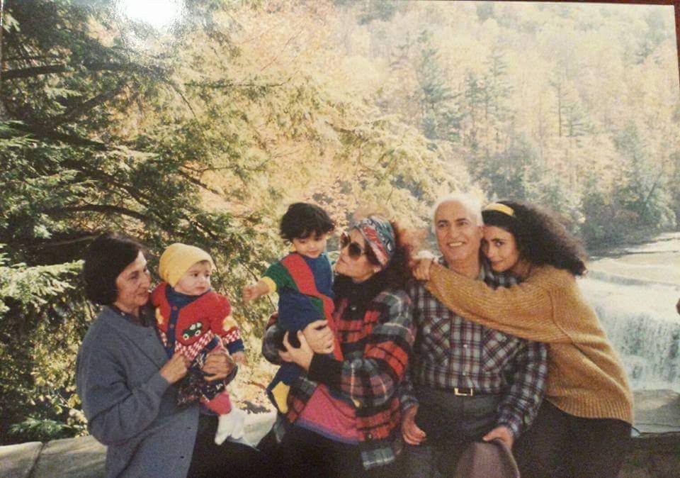 Family gathered outdoors by a river, smiling and holding children in a leafy park.