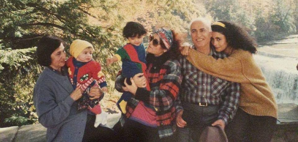 Four people sit outdoors on a blanket, laughing and hugging in a sunny park-like setting.