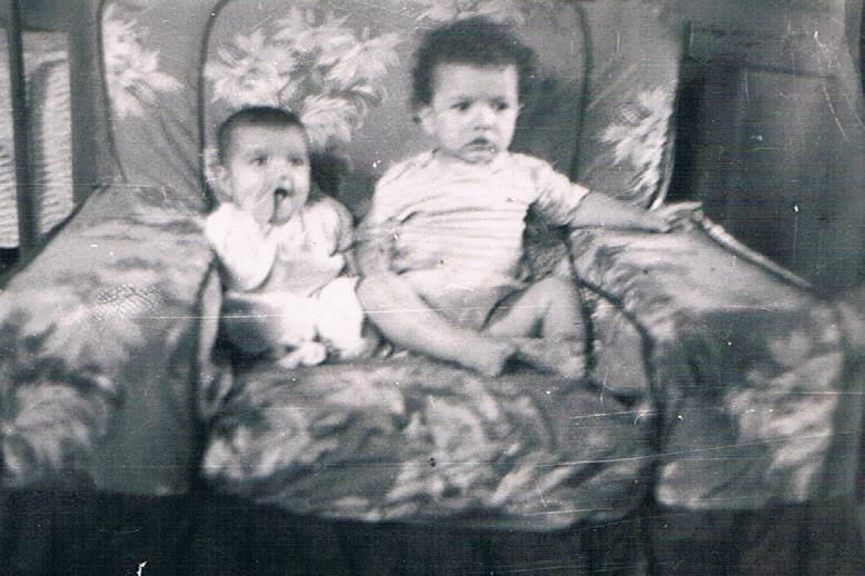 Two small children sitting on a patterned armchair, one sucking a finger, in a black-and-white photo