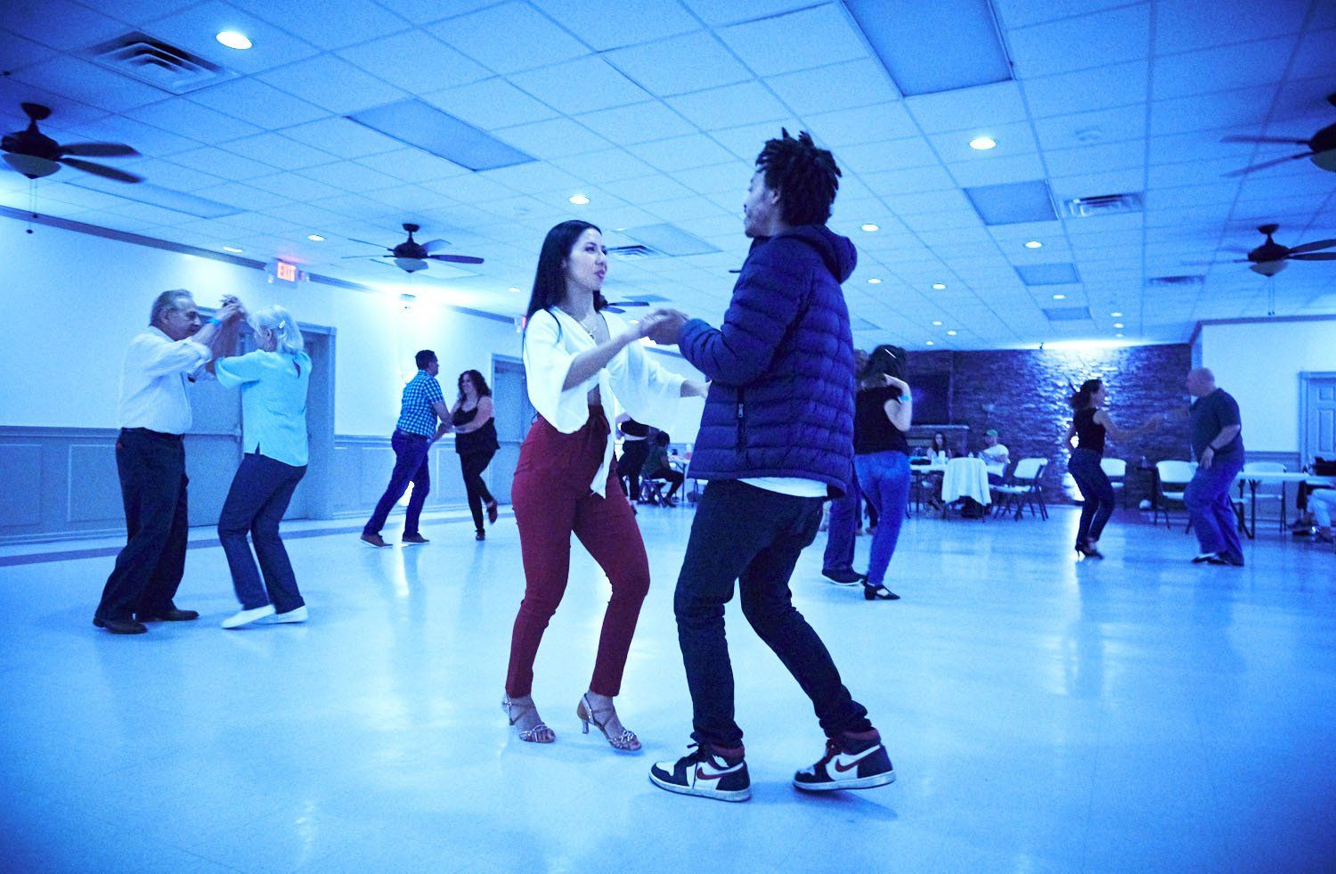 People dancing on a blue-lit indoor dance floor with a disco ball and ceiling lights.