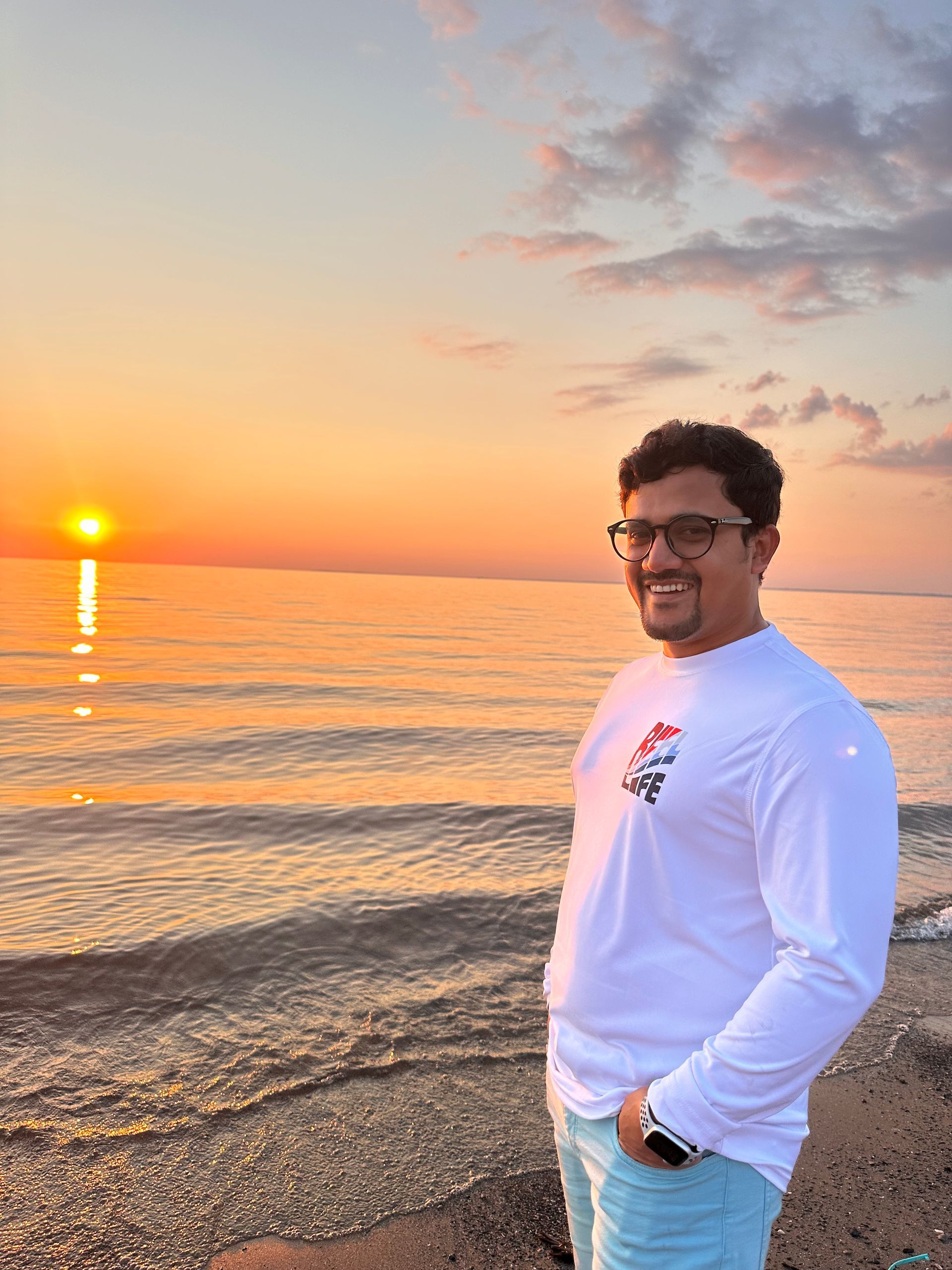 Person smiling on a pebble beach at sunset, with orange sky and calm water.