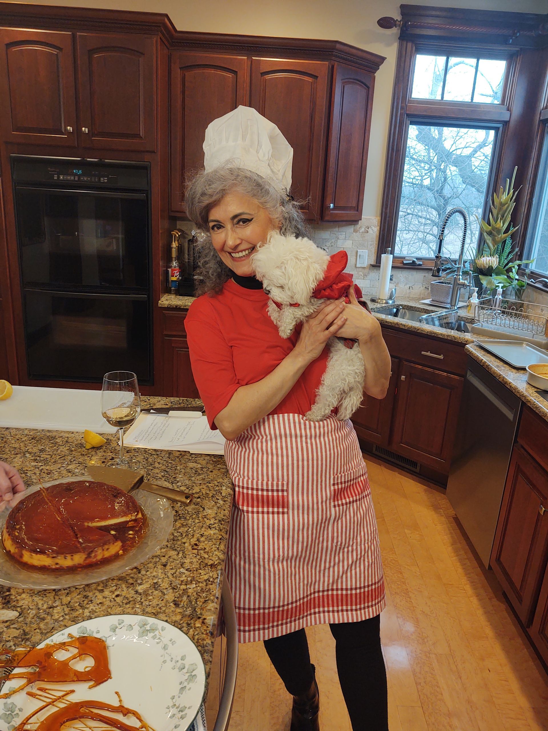 Woman in a kitchen holding a small white dog, wearing a red top, apron, and white chef hat.