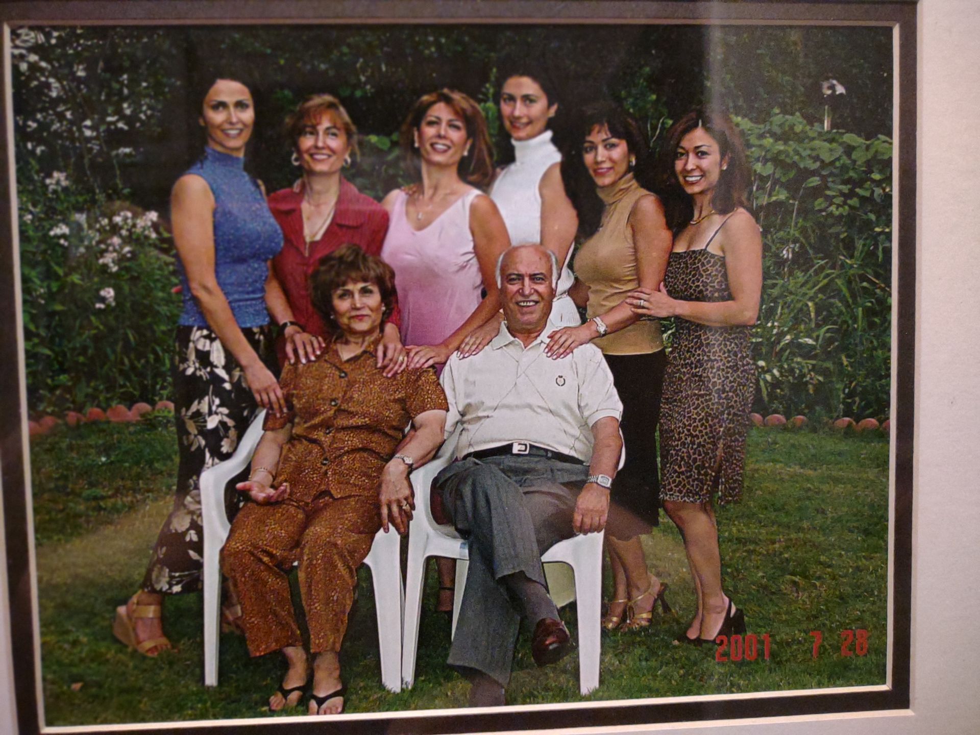 Group portrait of eight people outdoors, with two seated in white chairs and others standing behind them.
