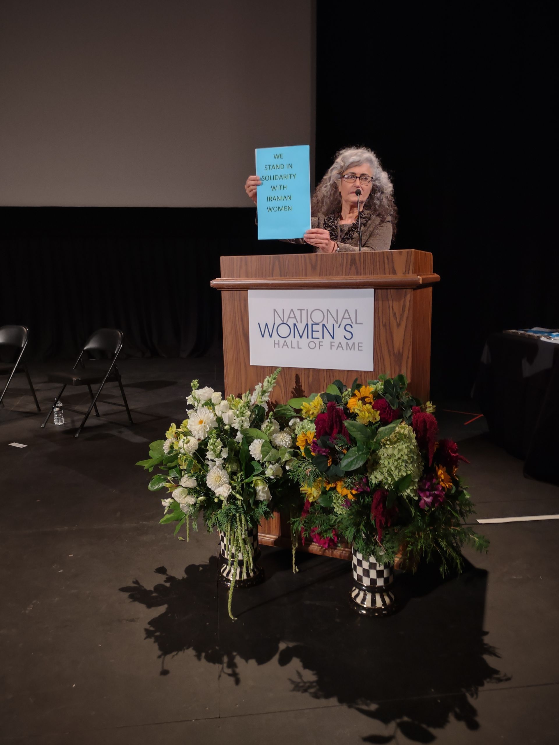 Woman at a podium holding a blue sign, with a large flower arrangement in front on a stage