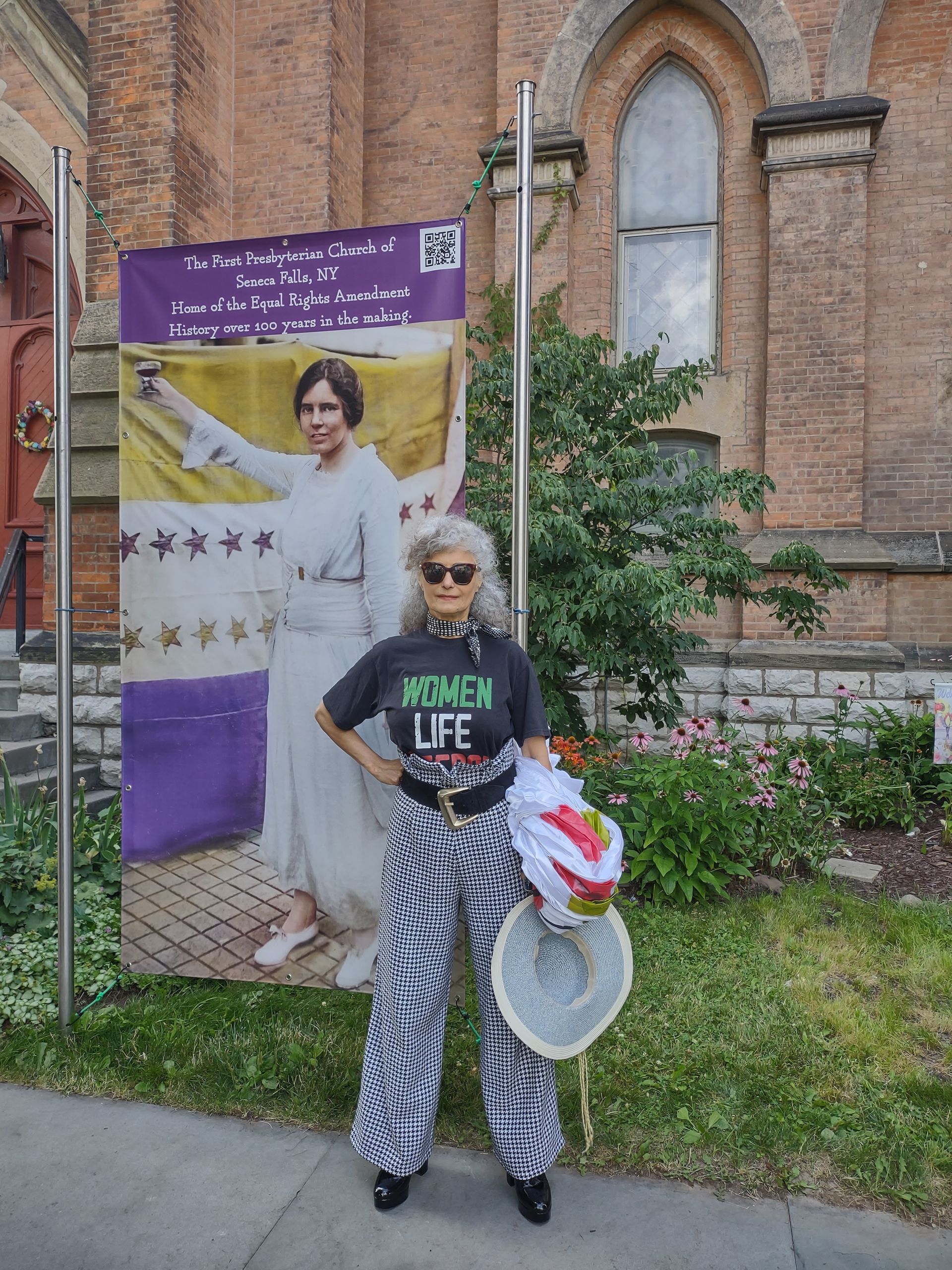 Person standing outside a brick building, holding a white basket and red flowers in front of a purple banner.