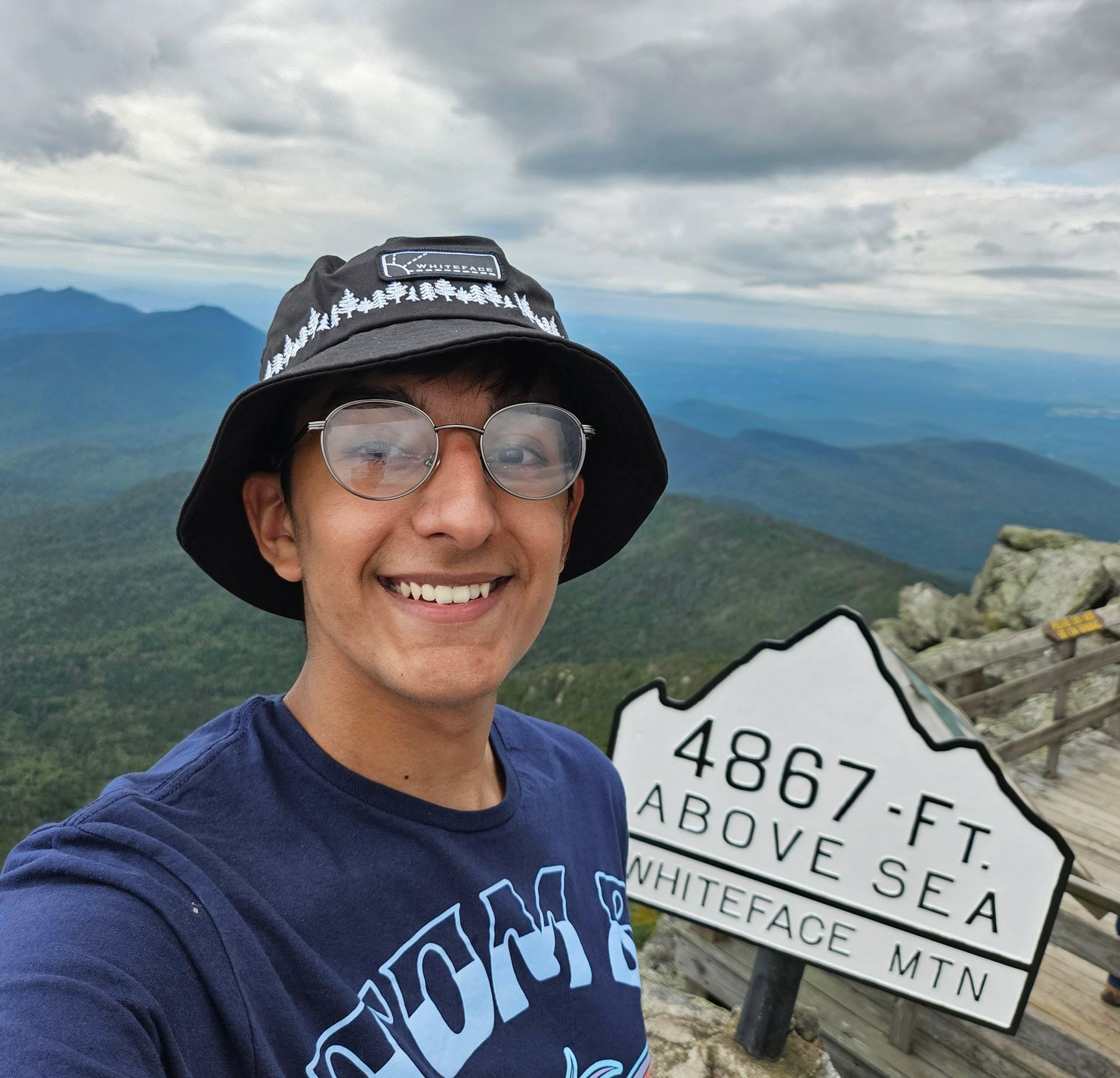 Smiling hiker selfie at Whiteface Mountain summit sign, cloudy mountain view in background