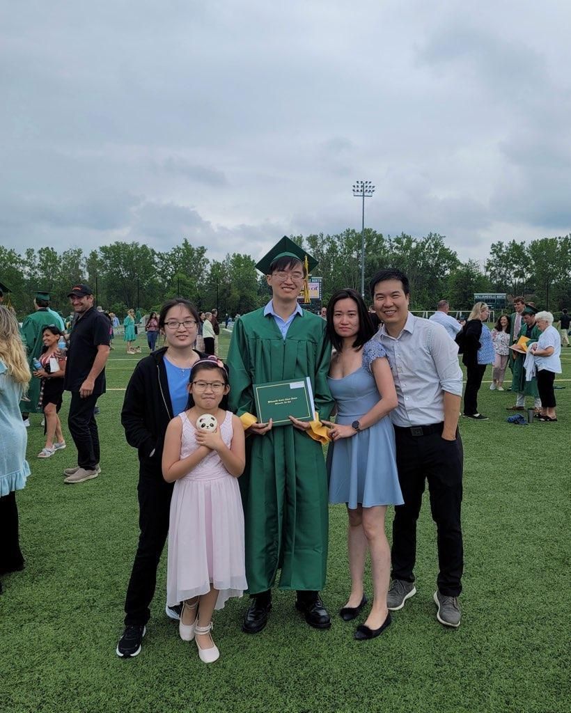 Family posing outdoors with a graduate in green cap and gown holding flowers on a grassy field.