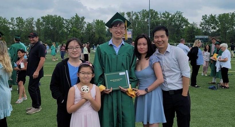 Group posing outdoors at a graduation ceremony, with a graduate in green cap and gown holding a diploma.