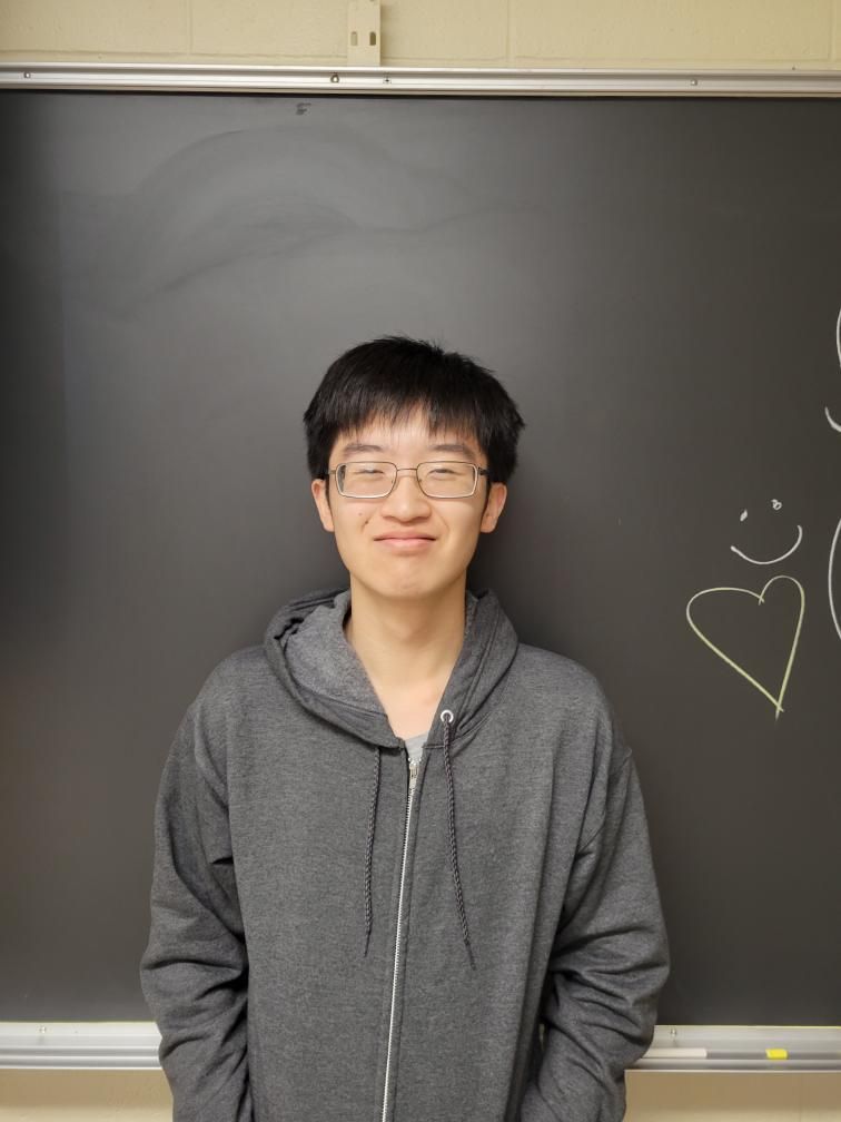 Person in a gray hoodie standing in front of a chalkboard, smiling, with a heart drawing on the right.
