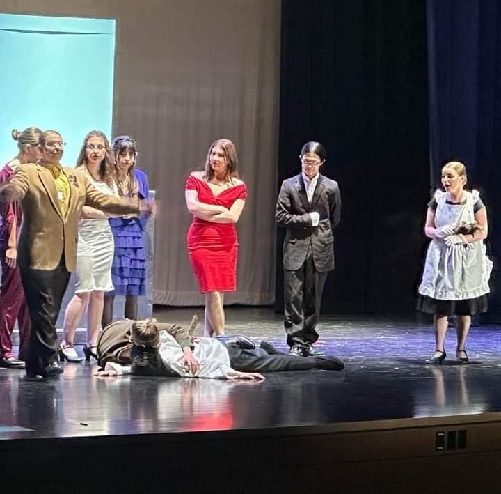Actors on a theater stage, with two performers lying on the floor and others standing nearby.