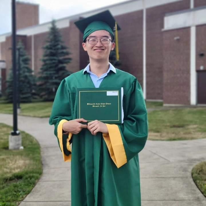Graduate in green cap and gown holding a diploma folder outdoors on a campus walkway