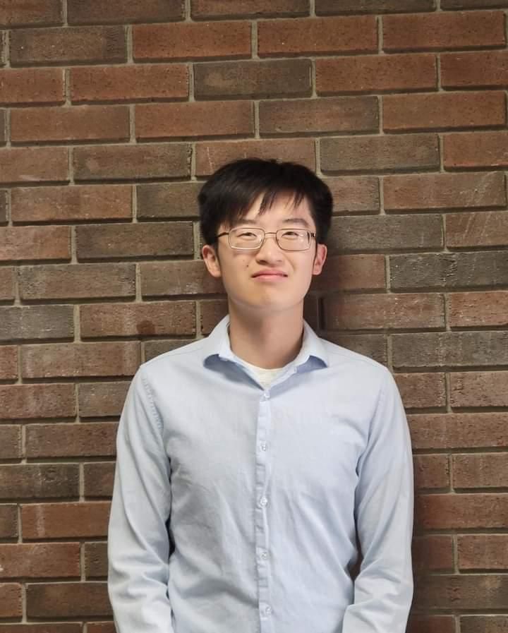 Person in a light blue shirt and glasses standing against a brick wall, smiling slightly.