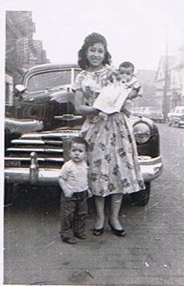 Woman in floral dress holding a baby, with a small child standing beside her in front of a vintage car.