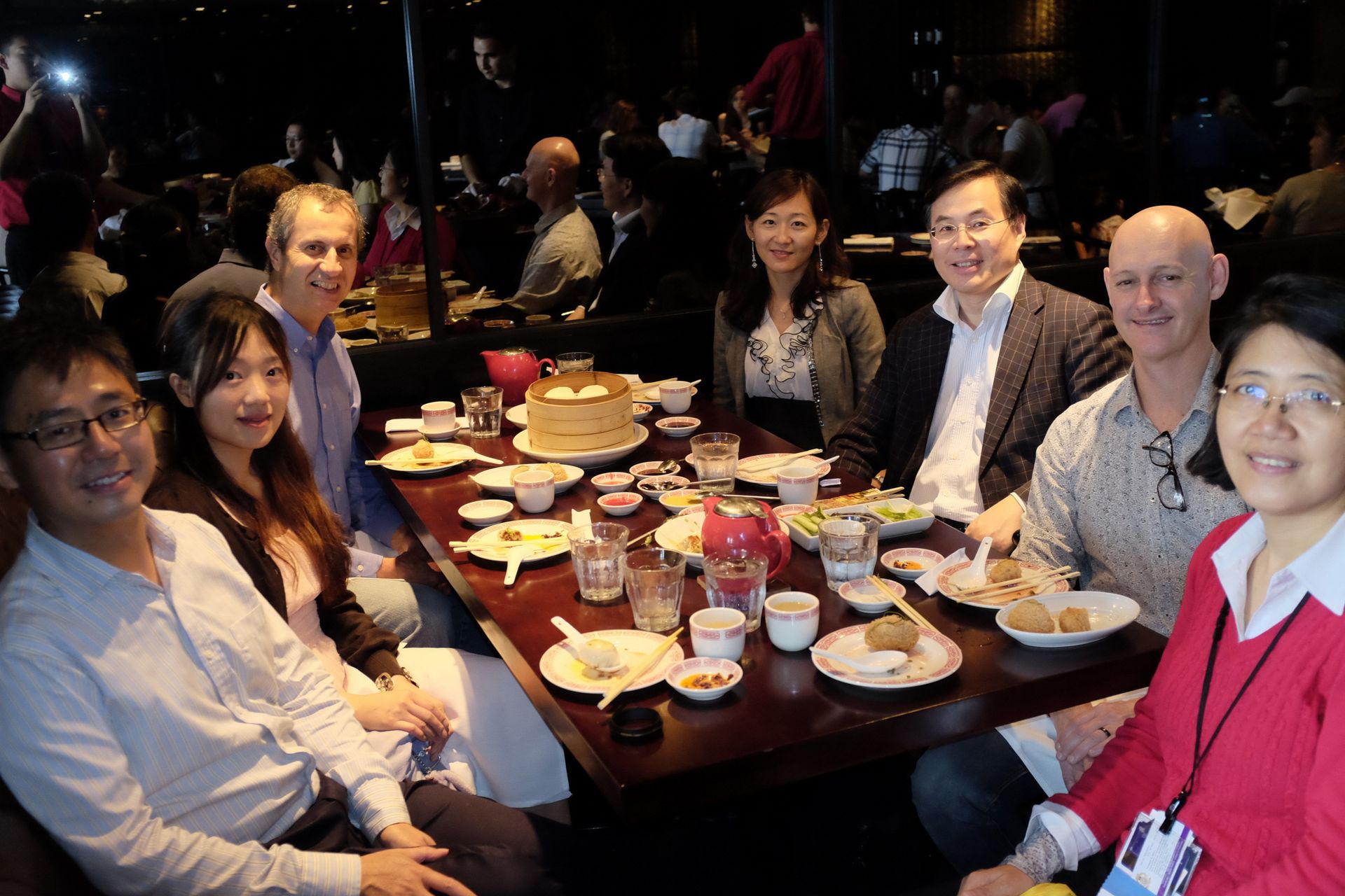 Group of people smiling around a restaurant table with drinks and dishes, posing for a meal together