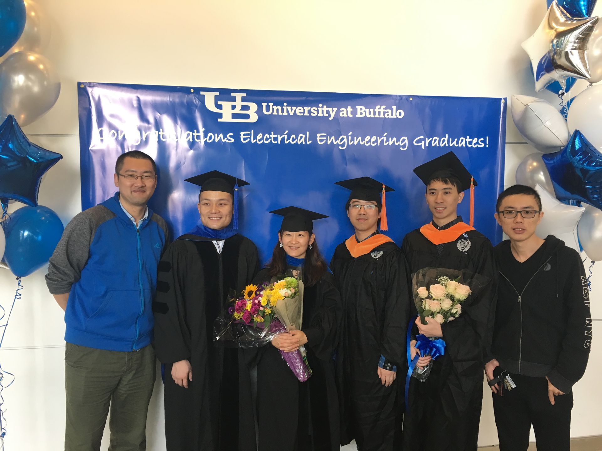 University at Buffalo graduates posing with flowers in front of a blue celebratory banner.