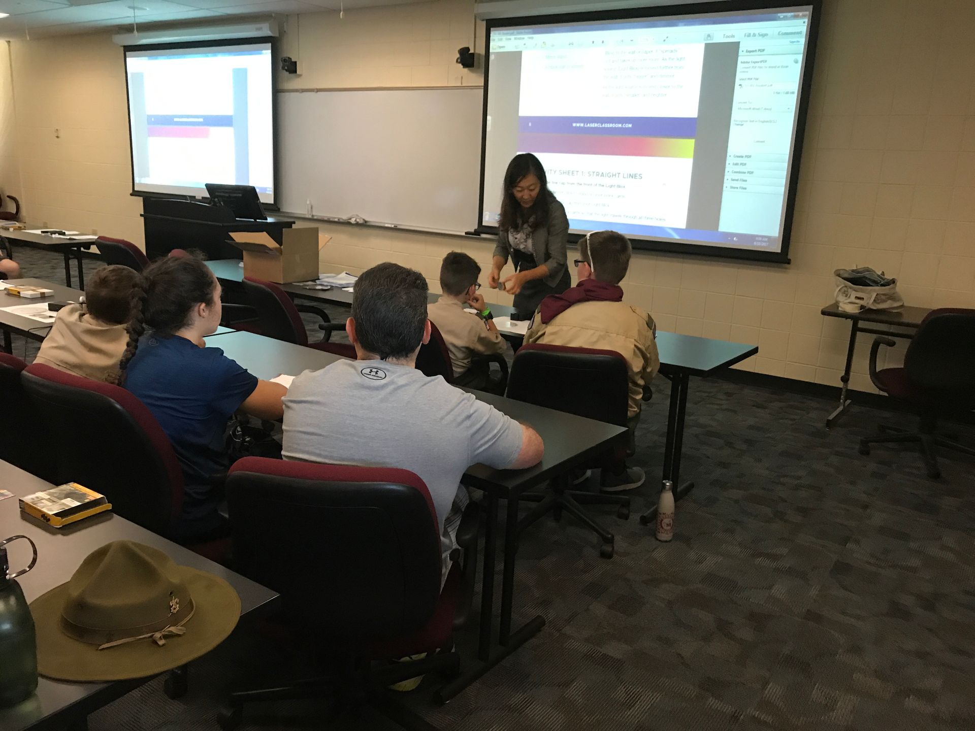 Classroom lecture with students watching a presenter at a desk, projected slides on two screens