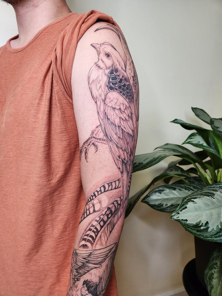 Arm with black floral and bird tattoo on a sleeveless peach shirt beside a leafy houseplant