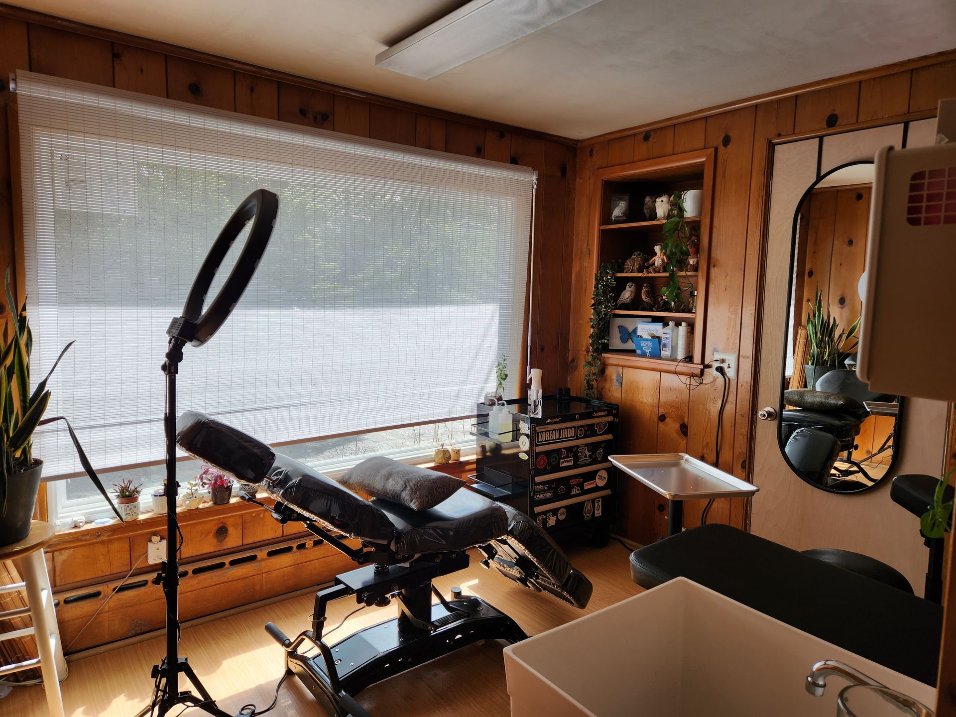 Wood-paneled room with a massage table, ring light, shelving, and a large sunlit window.