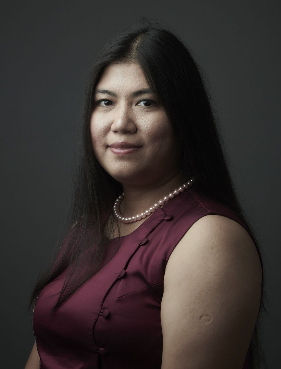 Portrait of a person in a maroon top and pearl necklace against a dark gray background